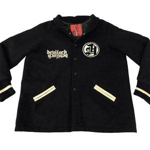 Puede incluir: Chaqueta universitaria negra con el texto "devilock yojiga" y un logotipo circular. La chaqueta tiene detalles en crema en los bolsillos, puños y cuello. La chaqueta está abotonada y tiene un forro acolchado.