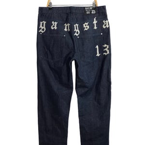 Puede incluir: Vaqueros de mezclilla azul oscuro con letras bordadas en blanco en la parte posterior que dicen "Gangsta 13".