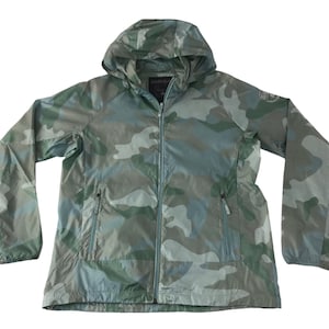 Könnte beinhalten: Eine Kapuzenjacke in Camouflage-Farben in Grün-, Grau- und Weißtönen. Die Jacke hat einen durchgehenden Reißverschluss, zwei Vordertaschen und eine Kapuze mit Kordelzug. Die Ärmel sind lang und die Jacke scheint leicht zu sein.