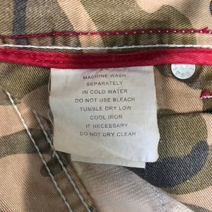 May include: A white care label with black text on a camouflage fabric background. The label reads "Machine wash separately in cold water. Do not use bleach. Tumble dry low. Cool iron if necessary. Do not dry clean."