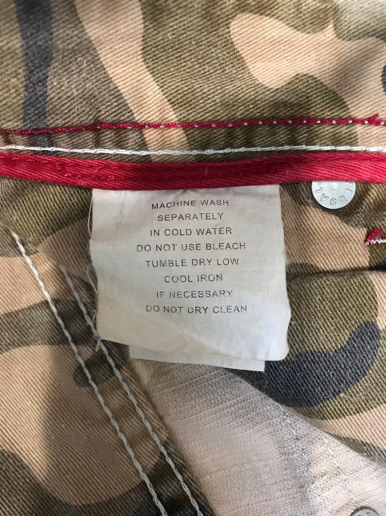 May include: A white care label with black text on a camouflage fabric background. The label reads "Machine wash separately in cold water. Do not use bleach. Tumble dry low. Cool iron if necessary. Do not dry clean."