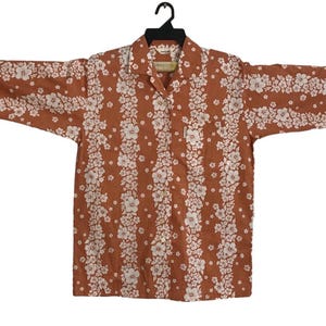 May include: A long-sleeved button-down shirt in a warm brown hue, adorned with vertical stripes of white floral patterns. The shirt features a classic collar and is displayed on a black hanger against a white background. The shirt's design is reminiscent of a Hawaiian shirt.