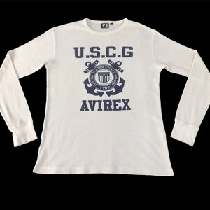 Könnte beinhalten: Cremefarbenes Langarmshirt mit gerippter Struktur. Das Shirt zeigt eine marineblaue Grafik mit dem Text "U.S.C.G" über einem Ankeremblem und dem Wort "AVIREX" darunter. Es hat einen Rundhalsausschnitt und eng anliegende Manschetten.