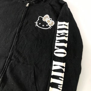 K&ouml;nnte beinhalten: Schwarzer Kapuzenpullover mit Rei&szlig;verschluss, der ein Hello Kitty-Motiv mit einer Leopardenmuster-Schleife auf der Brust zeigt. Der rechte &Auml;rmel zeigt die Worte "HELLO KITTY" in wei&szlig;en Blockbuchstaben. Der Hoodie hat eine Kapuze und einen Rei&szlig;verschluss.