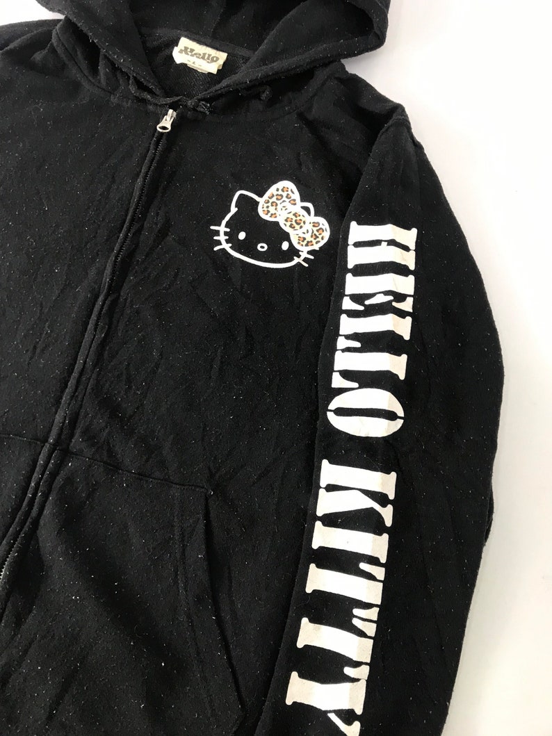 K&ouml;nnte beinhalten: Schwarzer Kapuzenpullover mit Rei&szlig;verschluss, der ein Hello Kitty-Motiv mit einer Leopardenmuster-Schleife auf der Brust zeigt. Der rechte &Auml;rmel zeigt die Worte "HELLO KITTY" in wei&szlig;en Blockbuchstaben. Der Hoodie hat eine Kapuze und einen Rei&szlig;verschluss.