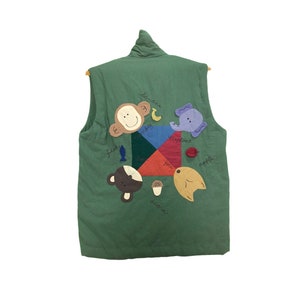 May include: A green quilted vest with a colourful geometric design, featuring appliquéd animal and food illustrations. The design includes a monkey, elephant, fish, acorn, and apple. The vest is sleeveless and has a high collar.