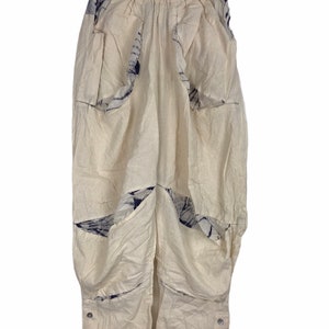 Könnte beinhalten: Weiße Leinenhose mit einem einzigartigen Design mit blauen und weißen gemusterten Stoffeinsätzen. Die Hose hat eine weite Beinweite und eine lockere Passform. Der Saum der Hose hat eine Reihe silberner Knöpfe.
