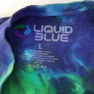 Puede incluir: Una camiseta tie-dye azul, verde y morada con una etiqueta blanca que dice "LIQUID BLUE" y "L" para el tama&ntilde;o. La etiqueta tambi&eacute;n incluye instrucciones de cuidado: 100% algod&oacute;n preencogido, lavar a m&aacute;quina con agua tibia, no usar blanqueador, secar en secadora a temperatura media, no planchar la decoraci&oacute;n, no lavar en seco, tejido en los EE. UU., ensamblado en M&eacute;xico, RH 81758.