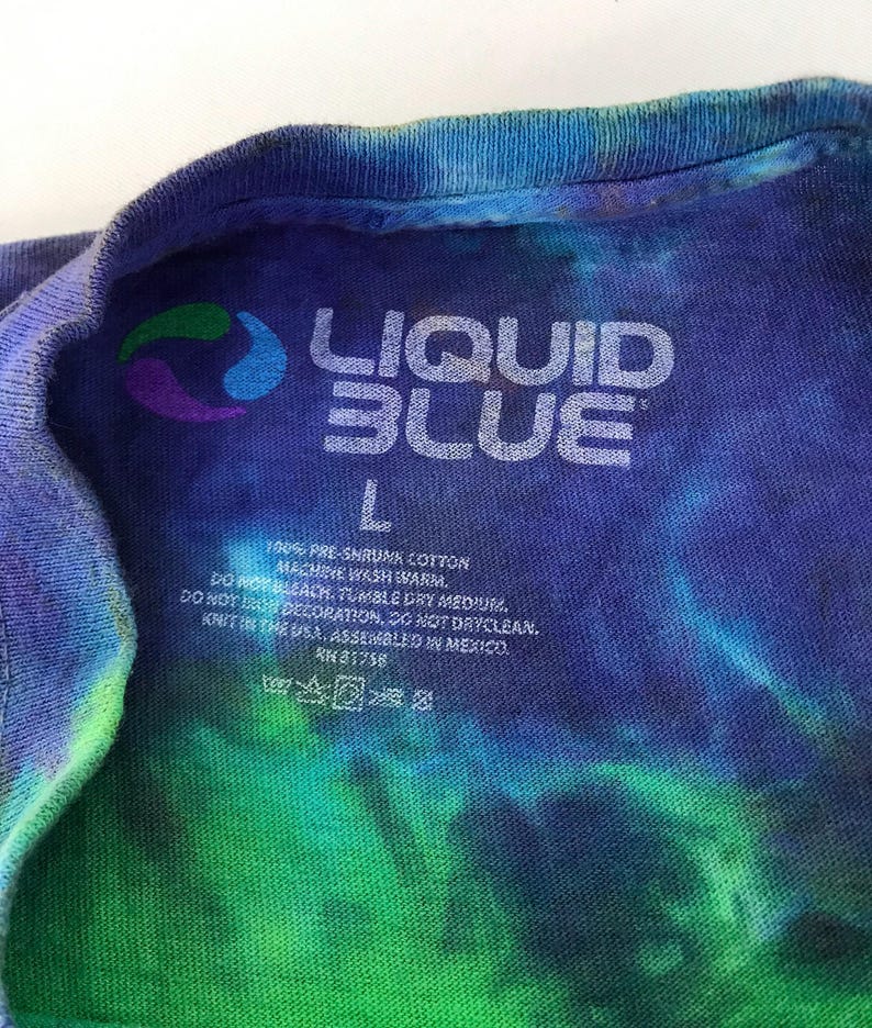 Puede incluir: Una camiseta tie-dye azul, verde y morada con una etiqueta blanca que dice "LIQUID BLUE" y "L" para el tama&ntilde;o. La etiqueta tambi&eacute;n incluye instrucciones de cuidado: 100% algod&oacute;n preencogido, lavar a m&aacute;quina con agua tibia, no usar blanqueador, secar en secadora a temperatura media, no planchar la decoraci&oacute;n, no lavar en seco, tejido en los EE. UU., ensamblado en M&eacute;xico, RH 81758.
