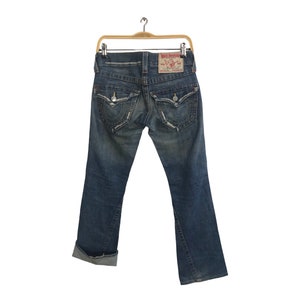 Könnte beinhalten: Blaue Jeans aus Denim mit Used-Details, Gesäßtaschen mit dekorativen Nähten und einem roten Lederpatch. Die Jeans werden auf einem Holzbügel vor weißem Hintergrund präsentiert.
