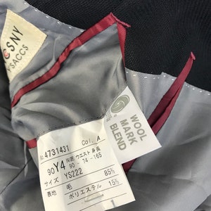 Puede incluir: Primer plano del interior de un blazer azul marino oscuro. El forro es gris claro con un ribete burdeos. Se ven varias etiquetas, incluida una etiqueta "WOOL MARK BLEND" y una etiqueta con texto y n&uacute;meros japoneses.