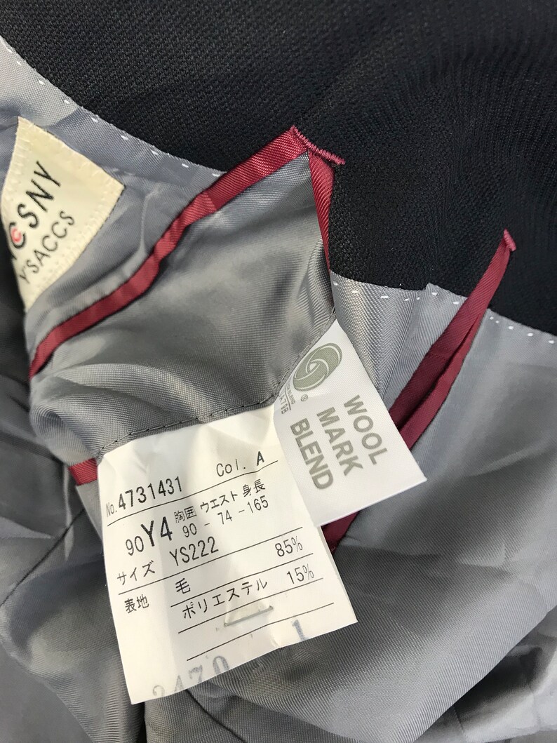 Puede incluir: Primer plano del interior de un blazer azul marino oscuro. El forro es gris claro con un ribete burdeos. Se ven varias etiquetas, incluida una etiqueta "WOOL MARK BLEND" y una etiqueta con texto y n&uacute;meros japoneses.