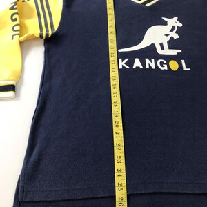 May include: Navy blue Kangol sweatshirt with yellow sleeves and a white kangaroo logo. The sleeves have black stripes and the word "NGOC" printed on them. The V-neck collar is white with navy and yellow trim. A measuring tape is next to the shirt.