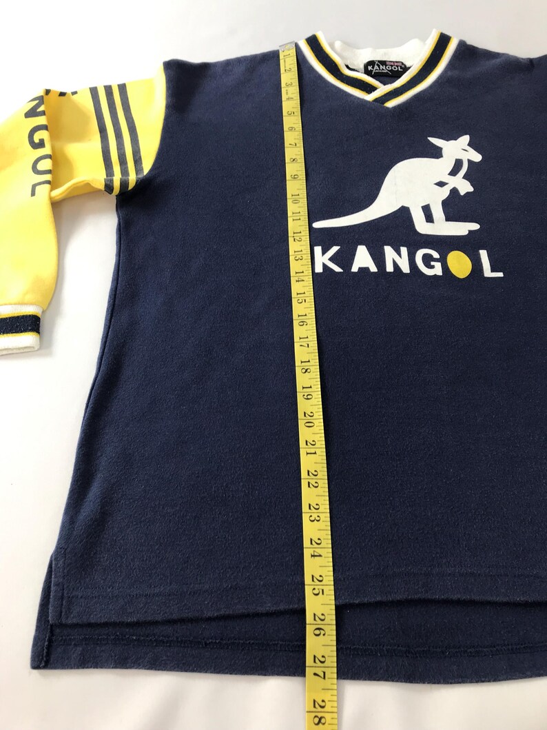 May include: Navy blue Kangol sweatshirt with yellow sleeves and a white kangaroo logo. The sleeves have black stripes and the word "NGOC" printed on them. The V-neck collar is white with navy and yellow trim. A measuring tape is next to the shirt.