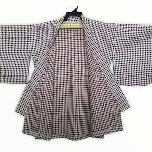 Op de afbeelding: Een wit en zwart geruite kimono met een rode en witte geruite voering. De kimono hangt aan een houten hanger.