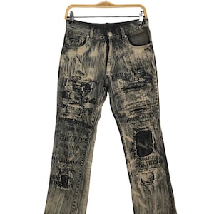 Könnte beinhalten: Verwaschene Jeans in Schwarz und Beige. Die Jeans hat Patches und aufgedruckte Textdetails. Sie wird auf einem Holzbügel vor weißem Hintergrund präsentiert.