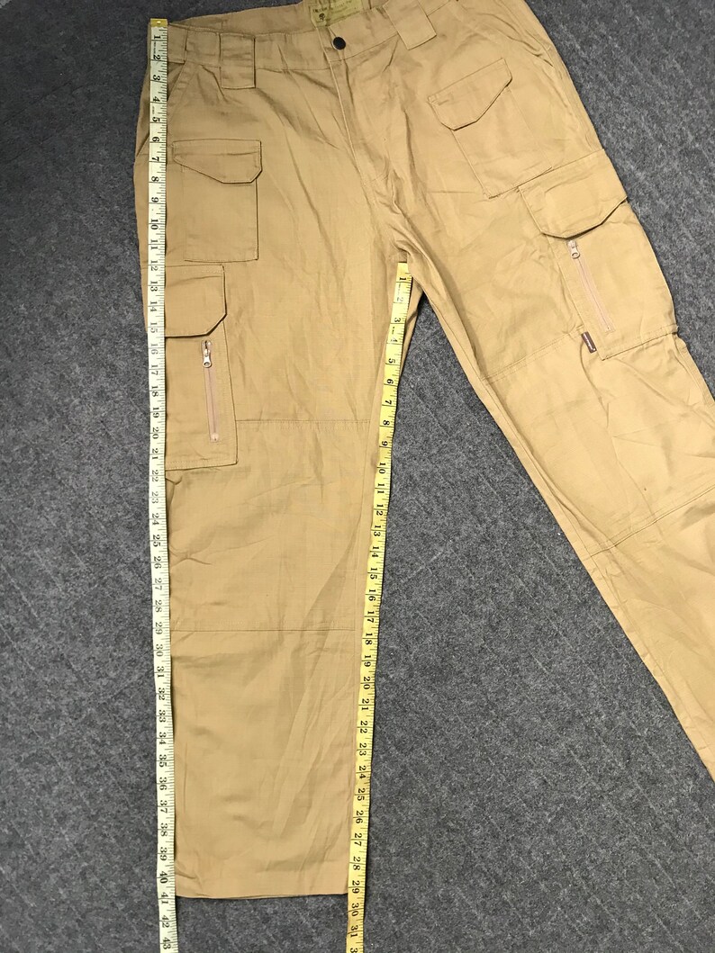 Pu&ograve; includere: Pantaloni cargo color cachi con pi&ugrave; tasche, tra cui alcune con cerniera. I pantaloni misurano circa 109 cm di lunghezza. I pantaloni hanno una chiusura con bottone e cerniera. Il tessuto sembra essere un materiale resistente e tessuto.
