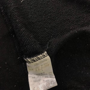 K&ouml;nnte beinhalten: Nahaufnahme eines schwarzen Kleidungsst&uuml;cketiketts mit japanischem Text und der Nummer SRD123079. Das Etikett enth&auml;lt auch Ma&szlig;e in Zentimetern und Details zur Stoffzusammensetzung, einschlie&szlig;lich Polyester und Baumwolle. Das Etikett lautet "MADE IN CHINA".