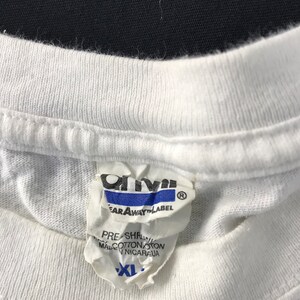 May include: Close-up of a white t-shirt collar with a tear-away label. The label is white with blue and black text, including the brand name "Anvil" and the size "XL". The shirt appears to be made of cotton.