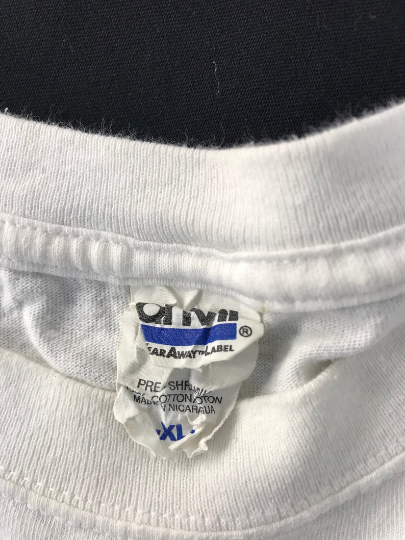 May include: Close-up of a white t-shirt collar with a tear-away label. The label is white with blue and black text, including the brand name "Anvil" and the size "XL". The shirt appears to be made of cotton.