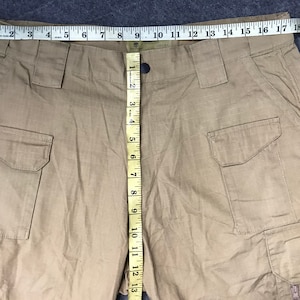 Pu&ograve; includere: Pantaloni cargo color kaki con pi&ugrave; tasche e chiusura con bottoni. I pantaloni sono misurati con un metro a nastro, che mostra una lunghezza di circa 33 cm. Il tessuto sembra essere un materiale resistente e tessuto.