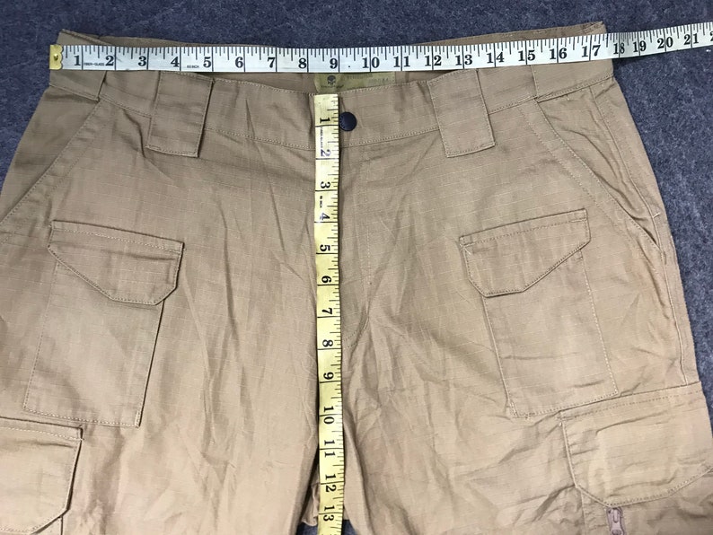 Pu&ograve; includere: Pantaloni cargo color kaki con pi&ugrave; tasche e chiusura con bottoni. I pantaloni sono misurati con un metro a nastro, che mostra una lunghezza di circa 33 cm. Il tessuto sembra essere un materiale resistente e tessuto.