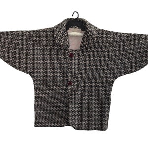 May include: A black and brown houndstooth jacket hangs on a black hanger. The jacket features a wide collar and three dark red buttons. The sleeves are wide and extend outwards. The jacket is made of a woven fabric.