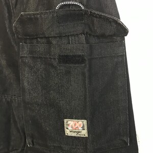 May include: Close-up of black denim trousers with a large cargo pocket and Velcro fastening. The pocket has a rectangular label with the text "700 Wrangler". White stitching detail is visible near the top.