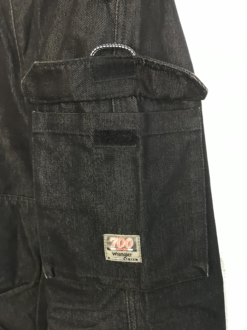 May include: Close-up of black denim trousers with a large cargo pocket and Velcro fastening. The pocket has a rectangular label with the text "700 Wrangler". White stitching detail is visible near the top.