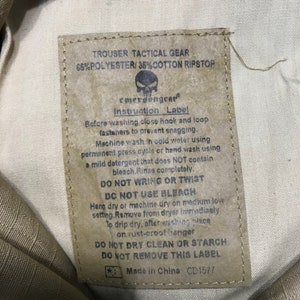 Pu&ograve; includere: Un'etichetta beige per pantaloni tattici con la scritta "TROUSER TACTICAL GEAR" e "65% POLYESTER/35% COTTON RIPSTOP". L'etichetta include istruzioni di lavaggio e il marchio "emersongear".