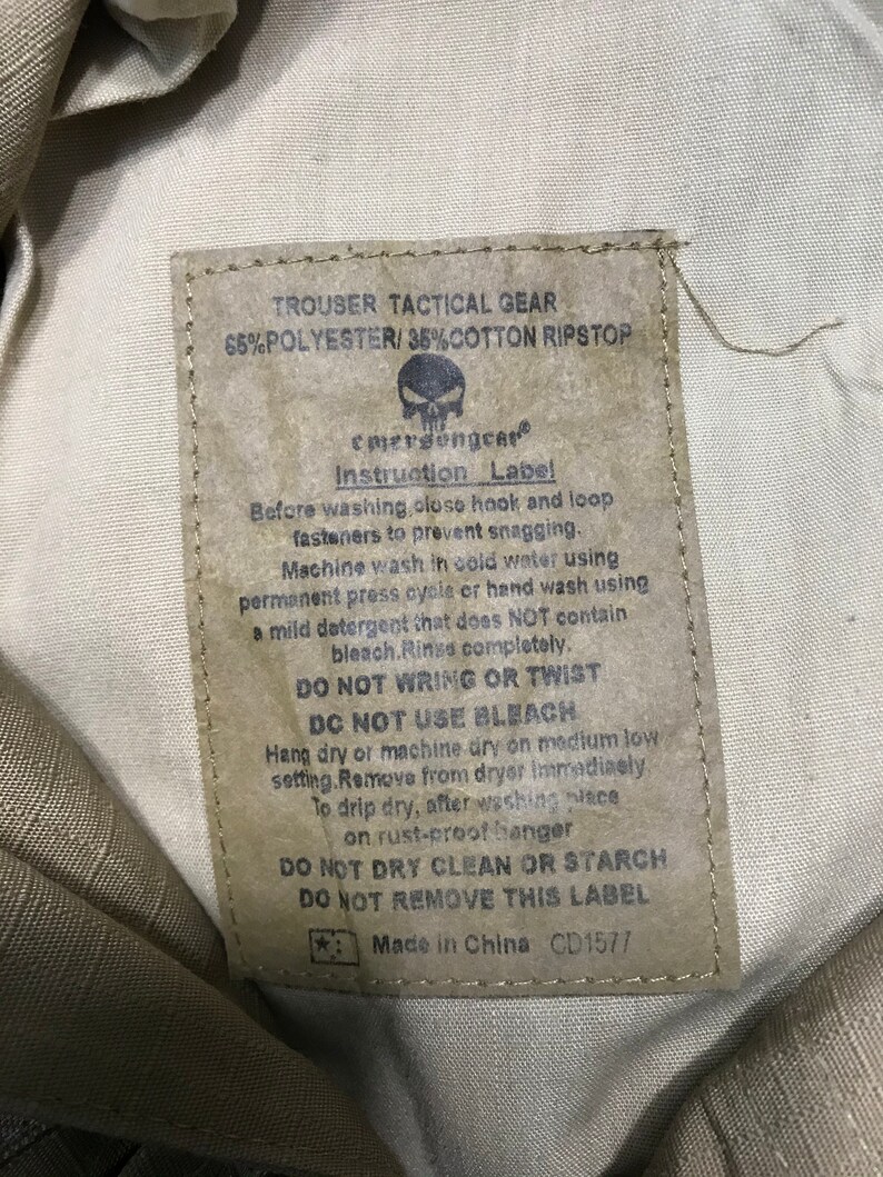 Pu&ograve; includere: Un'etichetta beige per pantaloni tattici con la scritta "TROUSER TACTICAL GEAR" e "65% POLYESTER/35% COTTON RIPSTOP". L'etichetta include istruzioni di lavaggio e il marchio "emersongear".