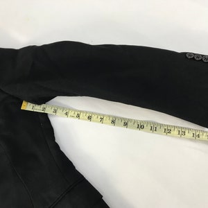 Puede incluir: Manga de blazer negra con una cinta m&eacute;trica amarilla que muestra una longitud de aproximadamente 40 cm. La manga tiene cuatro botones negros. El blazer es negro.