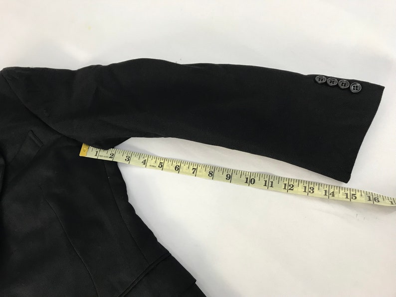 Puede incluir: Manga de blazer negra con una cinta m&eacute;trica amarilla que muestra una longitud de aproximadamente 40 cm. La manga tiene cuatro botones negros. El blazer es negro.