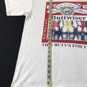 May include: White t-shirt with a graphic that reads "Buttwiser" in a red and white design, with the text "King of Rears" and "Las Vegas". The graphic features several rear views with colorful thongs. The shirt measures approximately 30 inches in length.