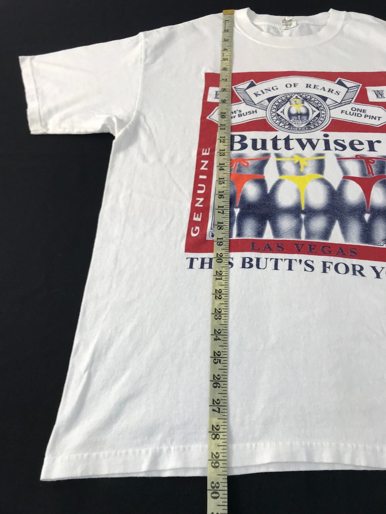 May include: White t-shirt with a graphic that reads "Buttwiser" in a red and white design, with the text "King of Rears" and "Las Vegas". The graphic features several rear views with colorful thongs. The shirt measures approximately 30 inches in length.