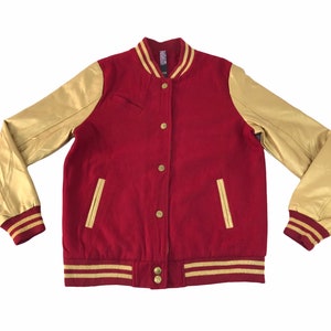 Puede incluir: Chaqueta universitaria roja y dorada con mangas doradas, cierre de botones y ribetes dorados en los puños y la cintura. La chaqueta tiene dos bolsillos delanteros y un cuello acanalado.