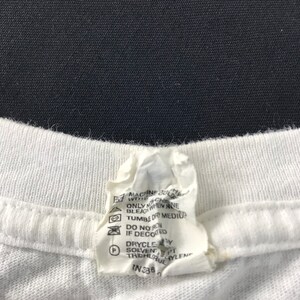 May include: Close-up of a white t-shirt collar with a care instruction tag. The tag includes washing symbols and text indicating machine wash, tumble dry medium, and dry clean instructions. The tag also has the number IN336.