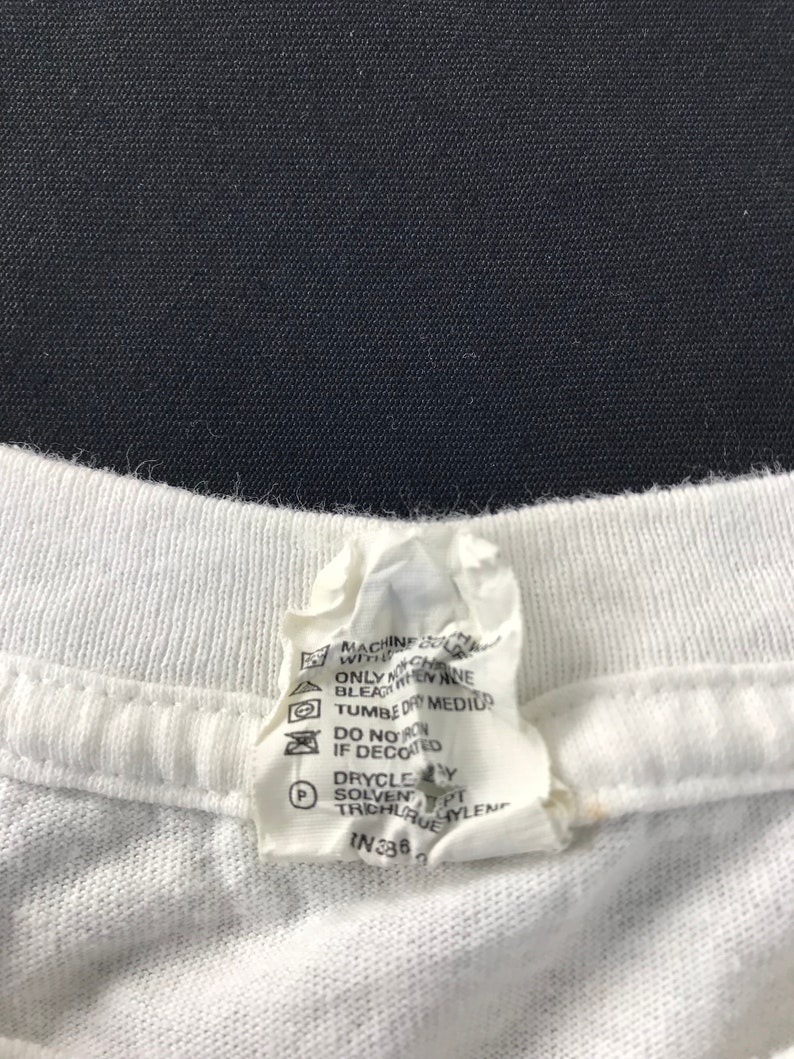May include: Close-up of a white t-shirt collar with a care instruction tag. The tag includes washing symbols and text indicating machine wash, tumble dry medium, and dry clean instructions. The tag also has the number IN336.