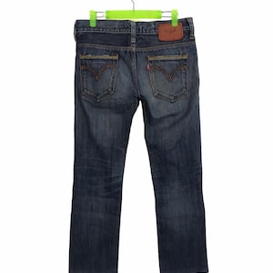 Könnte beinhalten: Dunkelblaue Jeans mit klassischem Fünf-Taschen-Design. Mit orangefarbenen Nähten, einem braunen Lederaufnäher und dem Levi's-Logo. Die Jeans hängen an einem grünen Kleiderbügel.