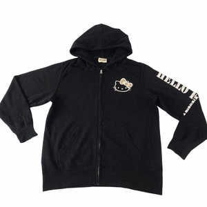 K&ouml;nnte beinhalten: Schwarzer Kapuzenpullover mit Rei&szlig;verschluss, der ein Hello Kitty-Motiv auf der Brust und den Schriftzug "HELLO KITTY" auf dem linken &Auml;rmel zeigt. Der Hoodie hat eine Kapuze und zwei Vordertaschen. Der Rei&szlig;verschluss ist silberfarben.