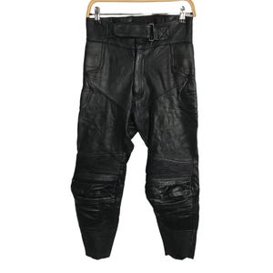 May include: Black leather motorcycle pants with a high waist and a belt. The pants have reinforced panels on the knees and thighs, and are hanging on a wooden hanger. The leather appears to be slightly worn.