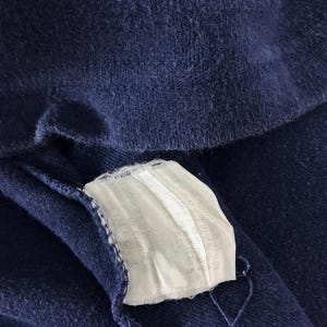 May include: Close-up of a navy blue fabric with a white tag attached. The tag is rectangular and has a ribbed texture. The fabric appears to be a soft knit material. The image is well-lit, highlighting the texture of the fabric.