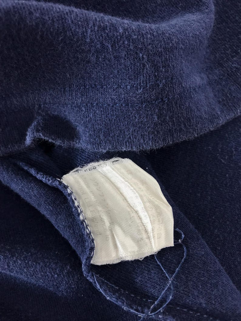 May include: Close-up of a navy blue fabric with a white tag attached. The tag is rectangular and has a ribbed texture. The fabric appears to be a soft knit material. The image is well-lit, highlighting the texture of the fabric.
