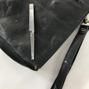 May include: Black leather clutch bag with a wrist strap and a silver metal tool. The bag has a zip closure and a textured surface. The silver tool is open and appears to be tweezers.