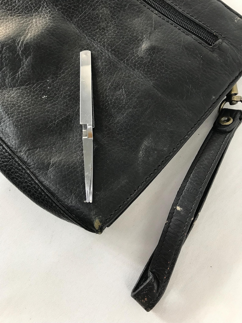 May include: Black leather clutch bag with a wrist strap and a silver metal tool. The bag has a zip closure and a textured surface. The silver tool is open and appears to be tweezers.