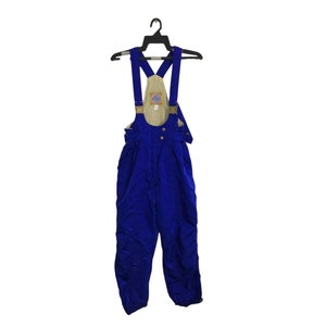 May include: Royal blue snow bib overalls hanging on a black hanger. The bib features tan fabric with a small patch and adjustable straps. The overalls have button closures and a relaxed fit, suitable for winter sports.