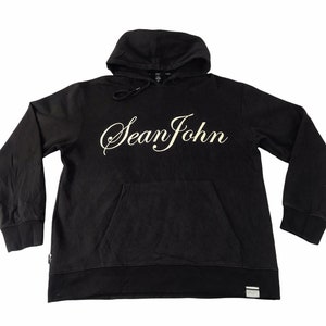 May include: Black hooded sweatshirt featuring a front pocket and the cursive text "Sean John" in white. The sweatshirt has long sleeves and a drawstring hood. A small tag is visible near the bottom hem.