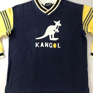 May include: Navy blue long-sleeve shirt with a white kangaroo logo and the word "KANGOL" in white. The shirt has yellow sleeves with black stripes and a white, black, and yellow trim around the V-neck collar.