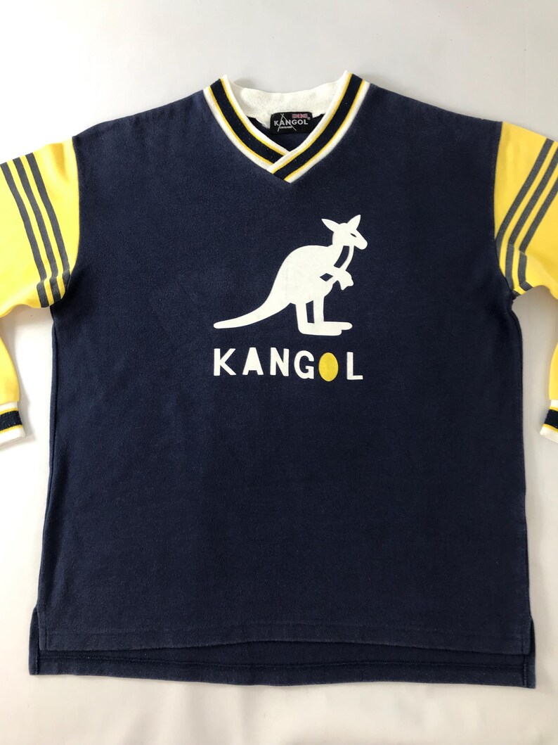 May include: Navy blue long-sleeve shirt with a white kangaroo logo and the word "KANGOL" in white. The shirt has yellow sleeves with black stripes and a white, black, and yellow trim around the V-neck collar.