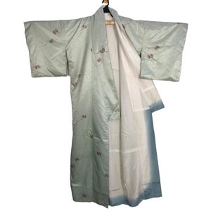 May include: A pale green kimono with a floral pattern and a white lining. The kimono is hanging on a wooden hanger. The sleeves are wide and the fabric has a subtle sheen.